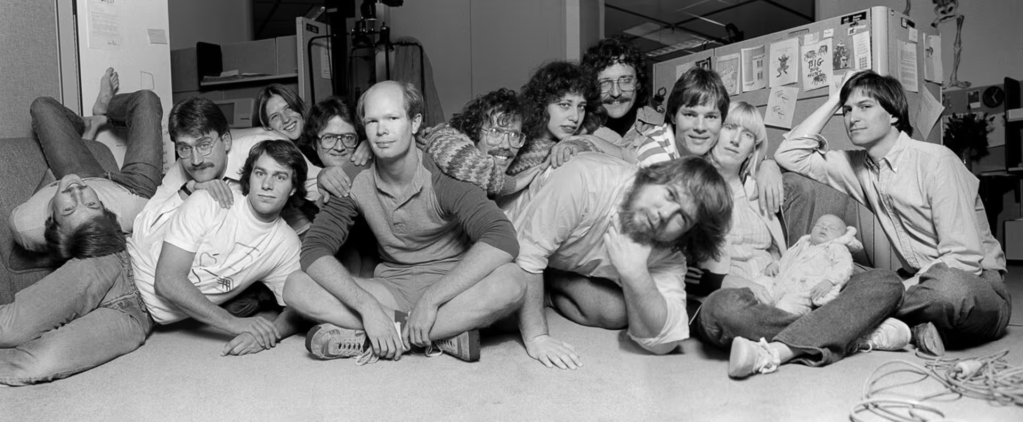 A group of people posing together in an office environment, showcasing a mix of relaxed and playful expressions. The scene captures a casual atmosphere, with individuals sitting and lying on the floor, surrounded by office equipment.