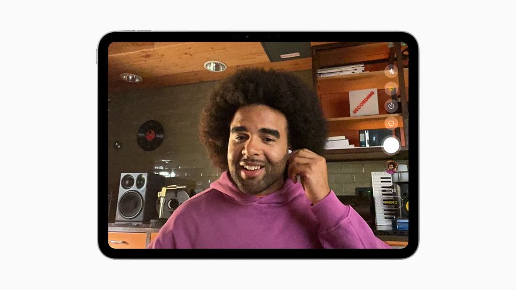 A person wearing AirPods and a pink hoodie smiles while using an iPad in a home setting with speakers and decor in the background.