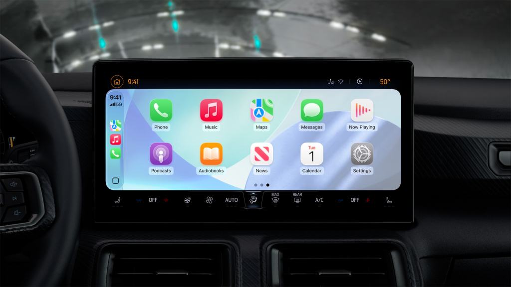 CarPlay 인터페이스가 표시된 자동차의 센터 디스플레이, iOS 26의 다양한 앱 아이콘과 시간, 온도 등의 정보가 포함되어 있음.