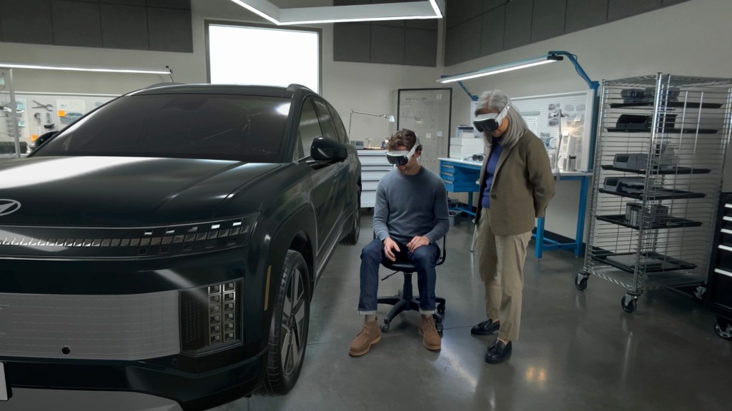 Two people wearing augmented reality headsets interact with a virtual vehicle in an indoor workspace.