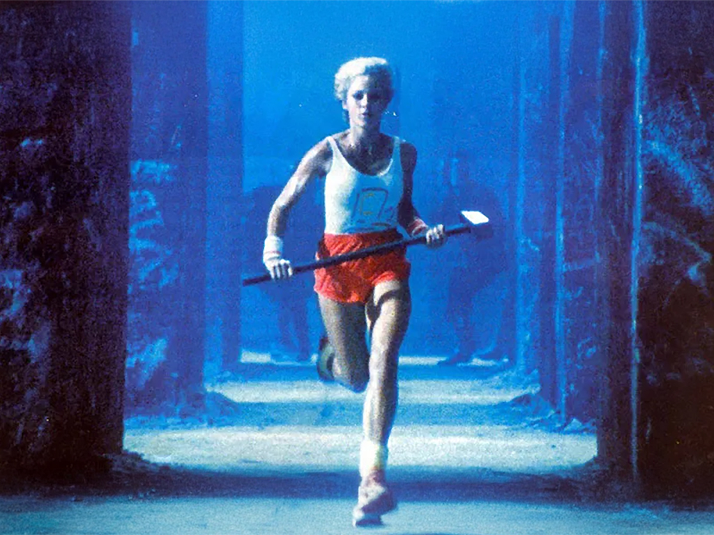 여성이 수중 환경에서 빨간 반바지와 흰색 상의를 입고 달리며 손에 도구를 들고 있는 모습.