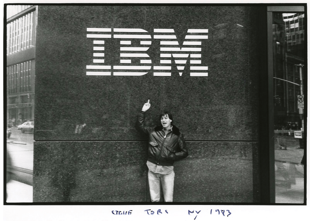 A person standing in front of an IBM building, pointing towards the IBM logo on the wall, dressed in a leather jacket and jeans, with city buildings in the background.