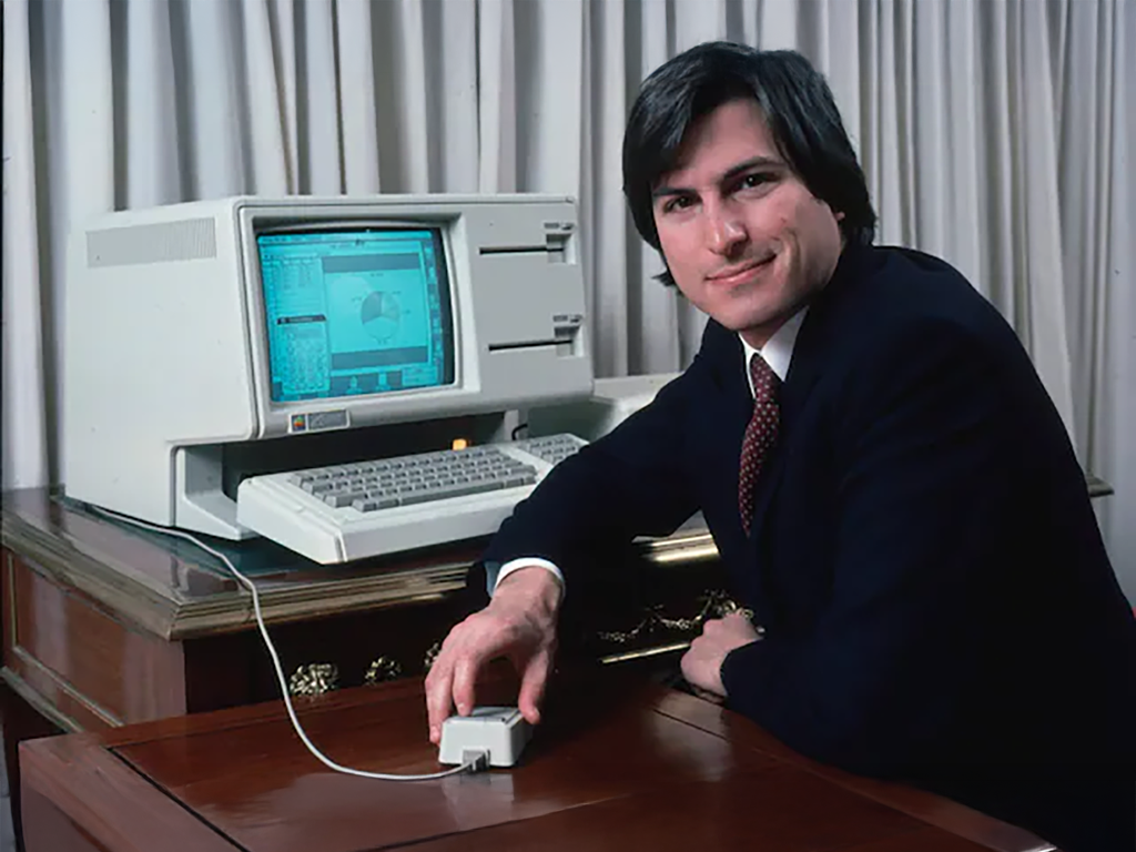 스티브 잡스가 컴퓨터 앞에 앉아 마우스를 조작하고 있는 모습. 배경에는 커다란 화면이 보이고, 고급스러운 테이블 위에 놓인 컴퓨터 시스템이 발견된다.