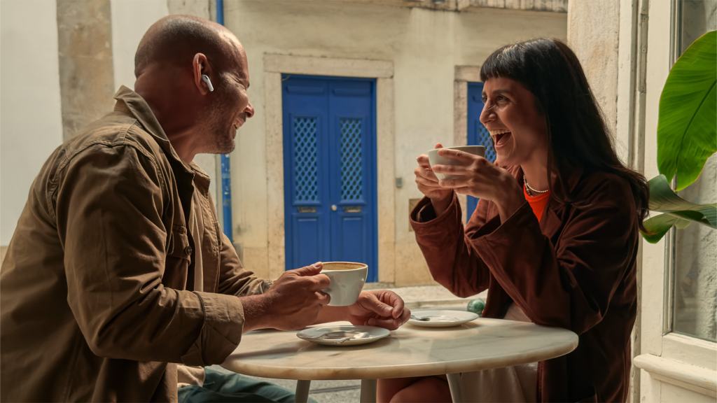 궁극의 청취 경험을 선사하는 AirPods Pro 3 공개 5 A man and a woman sitting at a café table, both smiling and enjoying conversation while wearing AirPods Pro. The background features blue doors and greenery, creating a vibrant atmosphere.