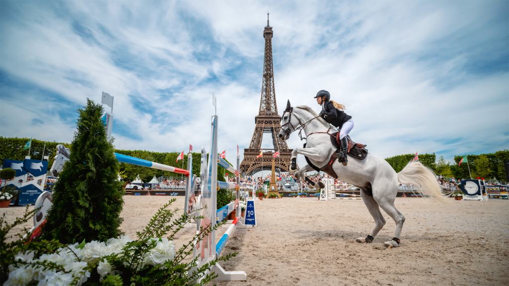 A rider on a white horse participating in a show jumping event in front of the Eiffel Tower, with decorative barriers and a blue sky in the background.