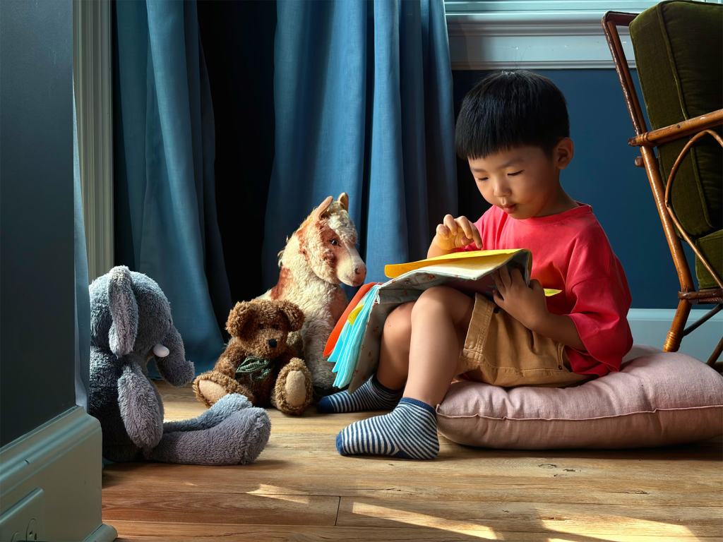 Apple, iPhone 17 발표 10 A young child sitting on a cushion, reading a book with stuffed animals nearby, surrounded by blue curtains and wooden flooring.