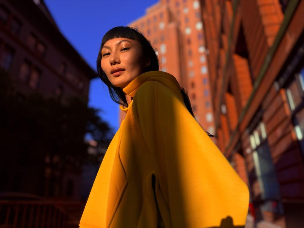 Apple, iPhone 17 Pro와 iPhone 17 Pro Max 발표 8 A woman in a yellow hoodie poses against a backdrop of buildings during sunset, showcasing warm light and shadows.
