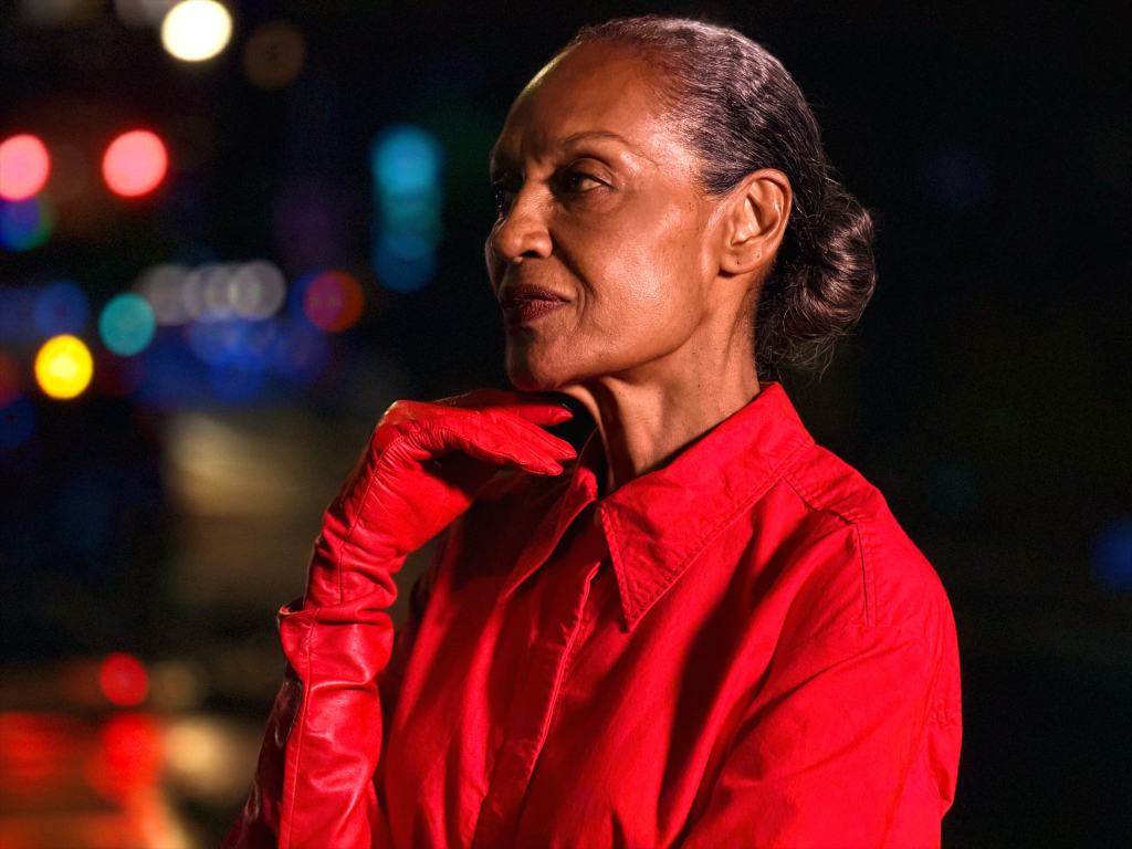 Apple, iPhone 17 Pro와 iPhone 17 Pro Max 발표 11 A woman in a bright red outfit with matching gloves poses thoughtfully in front of a blurred, illuminated background.
