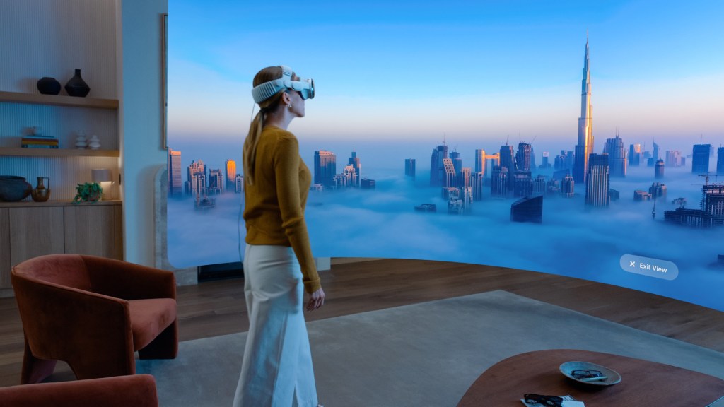 화면 앞에서 Apple Vision Pro를 착용하고 있는 여성. 뒤편에는 구름 위의 도시 풍경이 펼쳐져 있음.