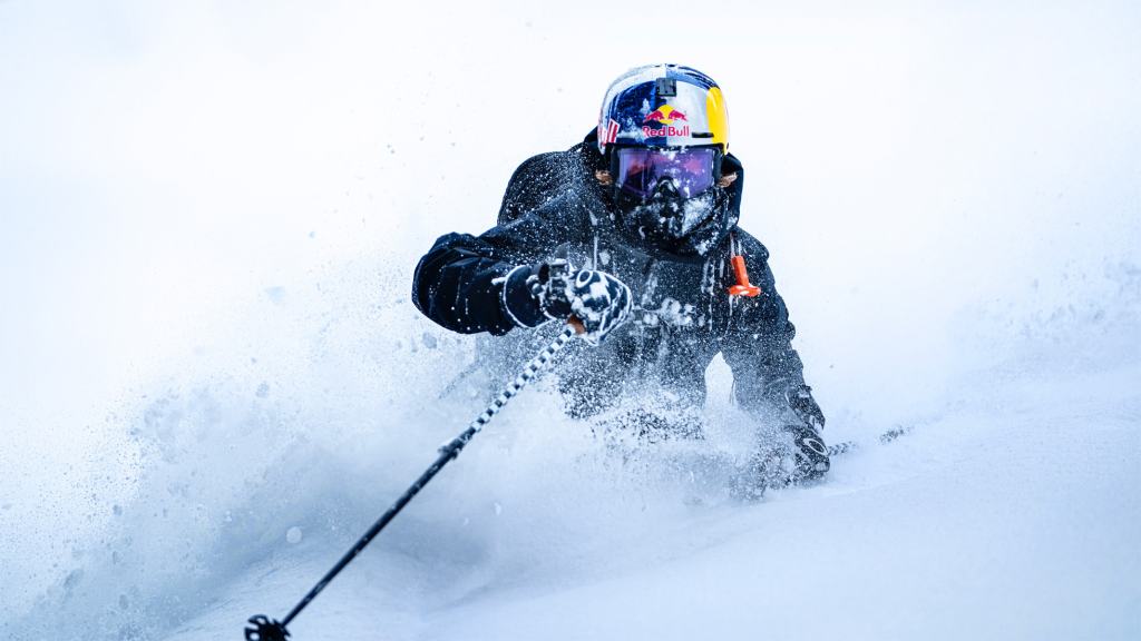 스키를 타고 있는 사람, 스노우 스프레이가 뒤로 튀어나오고 함박눈이 내리고 있는 환경에서 역동적인 동작을 보여줌.
