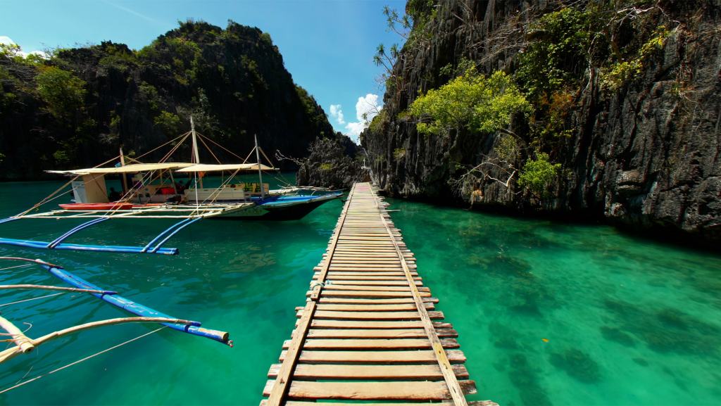 A wooden dock extends into clear turquoise water, with a boat moored alongside rocky cliffs and greenery in the background.