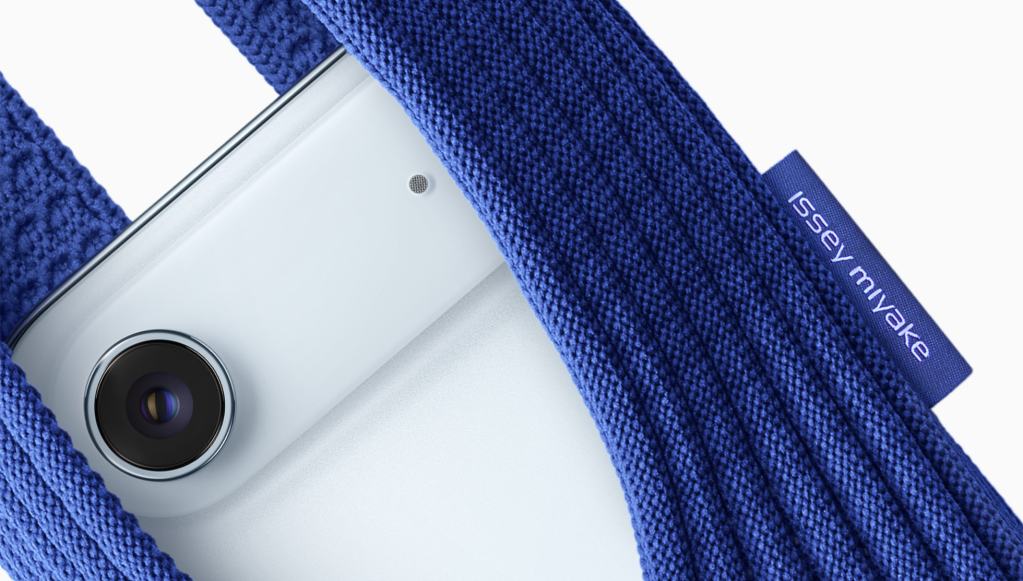 특징적인 푸른 색상의 ISSEY MIYAKE iPhone Pocket에 담긴 iPhone. 카메라 렌즈와 마이크 구멍이 보이며, 고유 로고 태그가 옆에 위치.