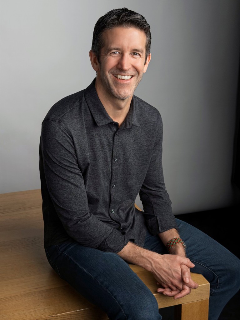 검은 셔츠를 입고 나무 테이블에 앉아 미소 짓고 있는 남성의 초상.