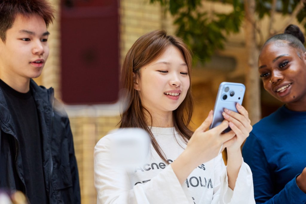 세 명의 젊은 사람들이 스마트폰을 보고 있는 모습, 웃고 있는 여성과 남성, 따뜻한 분위기.